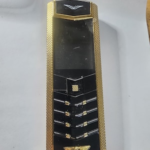 Può includere: Un telefono cellulare di lusso con una combinazione di colori nero e oro e una cornice dorata testurizzata. Il dispositivo include uno schermo nero, un pulsante quadrato centrale e una serie di tasti orizzontali. Un logo alato dorato è in basso.