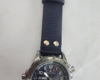 Może przedstawiać: Zegarek automatyczny Hamilton Khaki X-Wind z ciemnoniebieskim nylonowym paskiem z dwoma srebrnymi metalowymi nitami. Zegarek posiada czarną tarczę z białymi cyframi arabskimi, okienko datownika z dniem tygodnia oraz srebrne wskazówki.