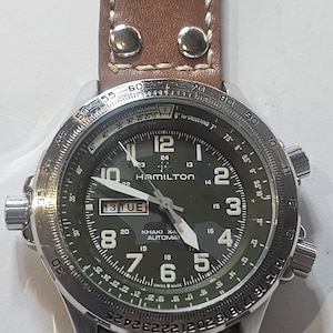 Puede incluir: Un reloj automático Hamilton Khaki X-Wind plateado con correa de cuero marrón. La esfera verde oliva muestra números y agujas blancas. La fecha es "13 TUE". El reloj tiene múltiples esferas y un bisel giratorio.