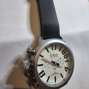 May include: A silver-toned U-BOAT watch with a black rubber strap. The watch face is off-white with black numerals and hands. The text "U-BOAT ITALO FONTANA" and "1001 METERS WATER RESISTANT" are visible on the watch face.