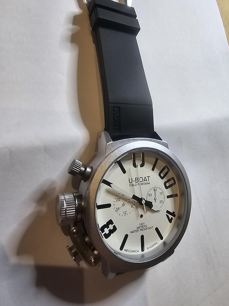 May include: A silver-toned U-BOAT watch with a black rubber strap. The watch face is off-white with black numerals and hands. The text "U-BOAT ITALO FONTANA" and "1001 METERS WATER RESISTANT" are visible on the watch face.