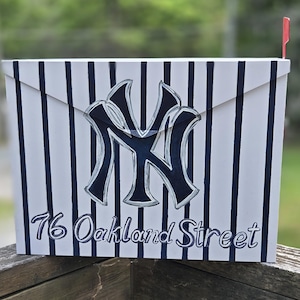 May include: A rectangular metal mailbox featuring a white base with vertical navy blue pinstripes. The front displays a large navy blue NY logo and the text 76 Oakland Street in a script font. A small red flag is attached to the right side.