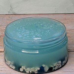 May include: A clear plastic jar filled with blue-tinted slime. The slime has small bubbles on the surface and is layered with small white and blue decorative stones at the bottom. The jar sits on a wooden surface.