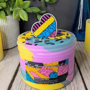 May include: A clear plastic container filled with multi-colored butter slime in yellow, blue, and purple. The top is decorated with black sprinkles and a heart-shaped topper. The label reads Wear Your Scars with Pride, Texture: Butter Slime, Scent: Almond, Strawberry, Indian Sandalwood, Jasmine, Orange Zest.