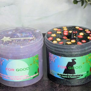 May include: Two clear plastic jars of slime. One is lavender with a silver star-shaped charm, the other is black with colorful flower-shaped decorations. Both jars have labels that say "FOR GOOD". The lavender jar's label says "Vanilla Bean & Pearls, Midnight Blackberry". The black jar's label says "Green Apple & Passion Fruit".