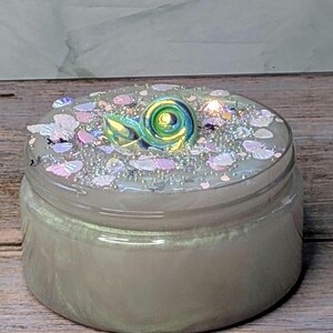 May include: A clear glass jar filled with a pearlescent, light green substance. The lid is adorned with iridescent sequins, small white beads, and a decorative, swirling, green and blue snail. The jar sits on a wooden surface.