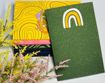 May include: A stack of notebooks with colorful covers. The top notebook is yellow with a mushroom design. A green notebook with a rainbow graphic is on the right. Pink and yellow floral sprigs are in the foreground.