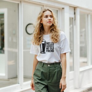 May include: White t-shirt with a black graphic of a beer and the text "HELLO DARKNESS MY OLD FRIEND". The shirt is paired with olive green pants. The image is taken outdoors.