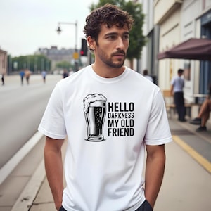 May include: White t-shirt with a black graphic of a beer glass and the text "HELLO DARKNESS MY OLD FRIEND". The beer glass is filled with dark beer and topped with a foamy head. The shirt is a classic crew neck.