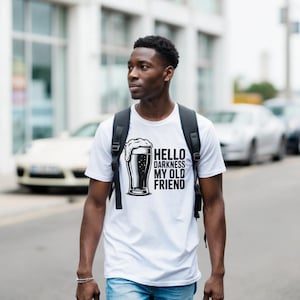 May include: White t-shirt with a black graphic of a beer glass and the text "Hello Darkness My Old Friend." The shirt is short-sleeved and worn by a person with a backpack.