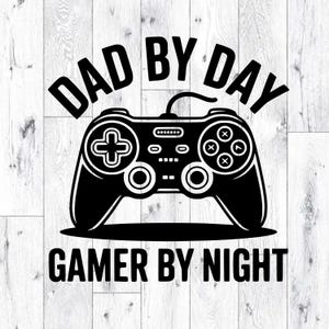 May include: Black and white graphic featuring a video game controller with the text "DAD BY DAY" above and "GAMER BY NIGHT" below. The controller has a cross-shaped directional pad, buttons, and joysticks.