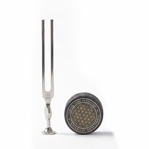 May include: A silver tuning fork with "512 Hz" etched on the base, next to a black tuning fork activator. The activator has gold text that reads "Tuning Fork Activator" around a gold geometric design.