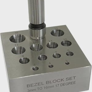 May include: A silver-colored bezel block set with a cylindrical punch tool. The block has 16 conical holes of varying sizes, ranging from 3mm to 16mm, and the text "BEZEL BLOCK SET 3mm TO 16mm 17 DEGREE" is visible on the side.