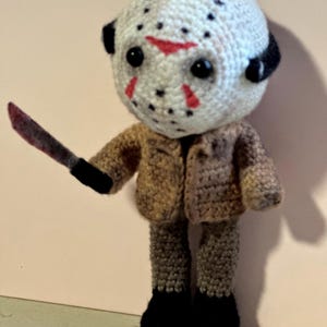 Könnte beinhalten: Handgemachte Häkelpuppe einer Figur mit weißer Hockeymaske mit roten Akzenten und schwarzem Haar. Die Puppe trägt eine braune Jacke und Hose, schwarze Stiefel und hält ein blutiges Messer. Die Puppe ist etwa 15 cm groß.