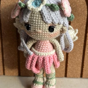 Könnte beinhalten: Eine handgefertigte Häkelpuppe in Form einer Fee. Die Puppe hat ein rosa Kleid, einen Blumenkranz und Flügel. Das Haar der Puppe ist hellviolett und die Details sind sorgfältig gearbeitet. Ein bezauberndes Beispiel handwerklichen Könnens.