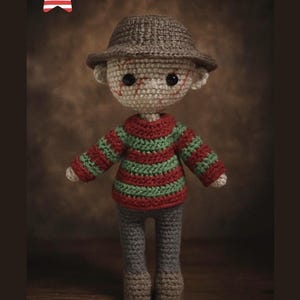 Könnte beinhalten: Gehäkelte Puppe einer Horrorfilmfigur, die einen gestreiften rot-grünen Pullover, eine graue Hose und einen braunen Hut trägt. Die Puppe hat aufgestickte Gesichtszüge und eine aufgestickte Narbe. Die Puppe ist etwa 20 cm groß.