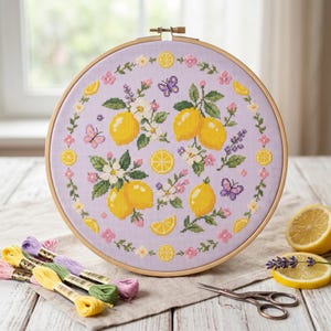 Puede incluir: Bordado de punto de cruz en un bastidor de madera. El diseño presenta limones amarillos, flores blancas, hojas verdes y mariposas moradas sobre un fondo lavanda. Hilos de bordar y tijeras en primer plano. Diámetro aproximado de 25 cm.