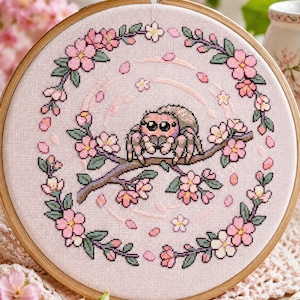 Op de afbeelding: Kruissteekborduurwerk in een houten hoepel. Het ontwerp toont een spin die op een tak met roze kersenbloesems zit. De achtergrond is een lichtroze stof en de hoepel is vastgezet met een messing sluiting.