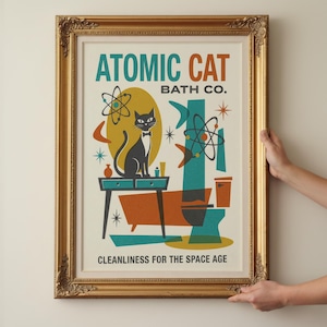 May include: A framed vintage-style print titled "Atomic Cat Bath Co." features a black cat in a bow tie, atomic symbols, and a retro bathroom scene in teal, orange, and yellow. The print is held by hands and framed in a gold ornate frame.