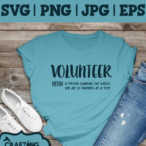 May include: A light blue t-shirt with the text "VOLUNTEER noun: a person changing the world one act of kindness at a time" printed on it. The shirt is laid out on a wooden surface with a pair of white sneakers and a pair of blue jeans.