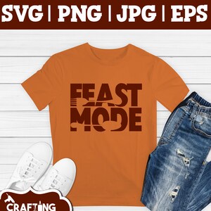 May include: Orange t-shirt with the text "Feast Mode" in brown letters. The letters are layered to create a design that includes a turkey and a fork. The t-shirt is on a wooden surface with a pair of white sneakers and blue jeans.