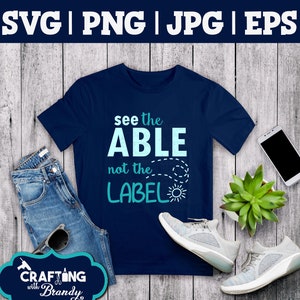 May include: A navy blue t-shirt with the text "see the ABLE not the LABEL" in a light blue color with a sun graphic. The t-shirt is laid out on a wooden surface with a pair of white sneakers, a pair of blue jeans, a cell phone, and a potted succulent plant.