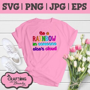 May include: A pink t-shirt with the text "Be a RAINBOW in someone else's cloud" in rainbow colors. The shirt is laid out on a white wooden surface with white sneakers and a bouquet of pink and white flowers.
