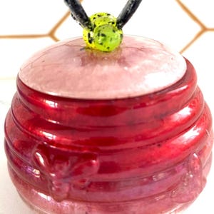 May include: A handcrafted, red ceramic jar with a lid, featuring a bee-shaped handle. The jar has a glossy finish and a ribbed design. The bee handle is yellow and black. The jar is likely used for storage or decoration.