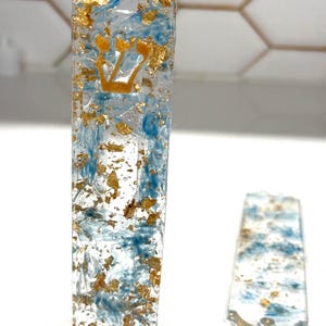 May include: A rectangular mezuzah case made of clear resin with embedded blue swirls and gold leaf flakes. The front features a gold-colored Hebrew letter Shin. A matching flat, rectangular resin piece with blue swirls and gold leaf lies nearby.