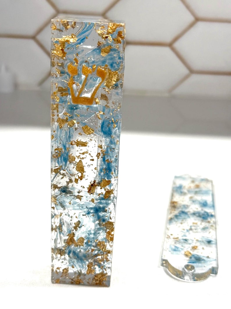 May include: A rectangular mezuzah case made of clear resin with embedded blue swirls and gold leaf flakes. The front features a gold-colored Hebrew letter Shin. A matching flat, rectangular resin piece with blue swirls and gold leaf lies nearby.