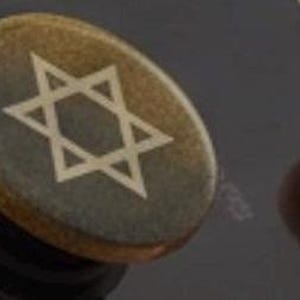 May include: A round, metallic phone grip featuring a Star of David design. The grip has a gold and bronze color scheme, with the star in a lighter shade. The grip is attached to a black phone.
