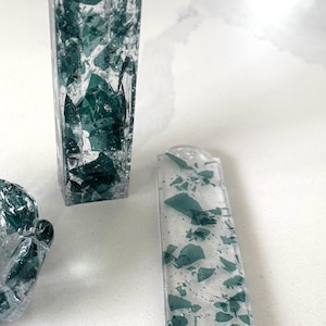May include: A vertical rectangular mezuzah case and a flat rectangular mezuzah case, both made of clear resin embedded with dark green glass shards. The vertical case features a silver Hebrew letter Shin on the front surface, while the flat case has rounded ends.
