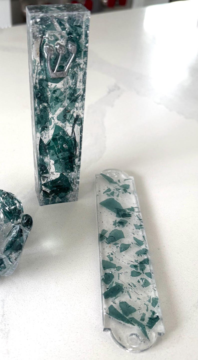 May include: A vertical rectangular mezuzah case and a flat rectangular mezuzah case, both made of clear resin embedded with dark green glass shards. The vertical case features a silver Hebrew letter Shin on the front surface, while the flat case has rounded ends.