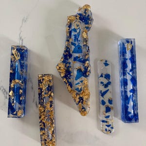 May include: A collection of five resin objects including three rectangular mezuzah cases with blue fragments and gold leaf, one map-shaped piece with blue and gold accents, and one flat, translucent white strip with blue and gold flecks.