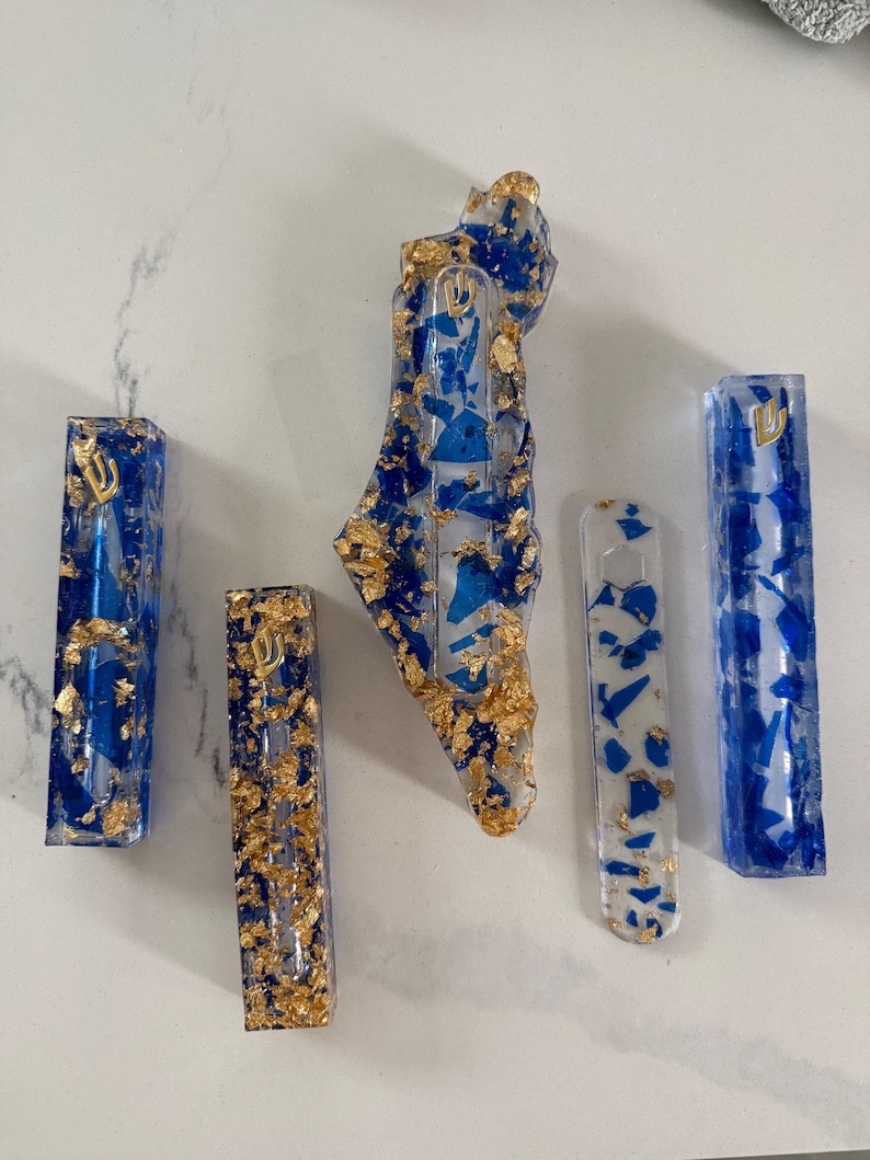 May include: A collection of five resin objects including three rectangular mezuzah cases with blue fragments and gold leaf, one map-shaped piece with blue and gold accents, and one flat, translucent white strip with blue and gold flecks.