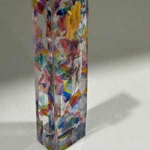 May include: A rectangular clear resin mezuzah case featuring an embedded mosaic of multicolored translucent shards in shades of pink, blue, yellow, and green, with a small, textured, light-colored Hebrew letter shin attached to the upper front surface.
