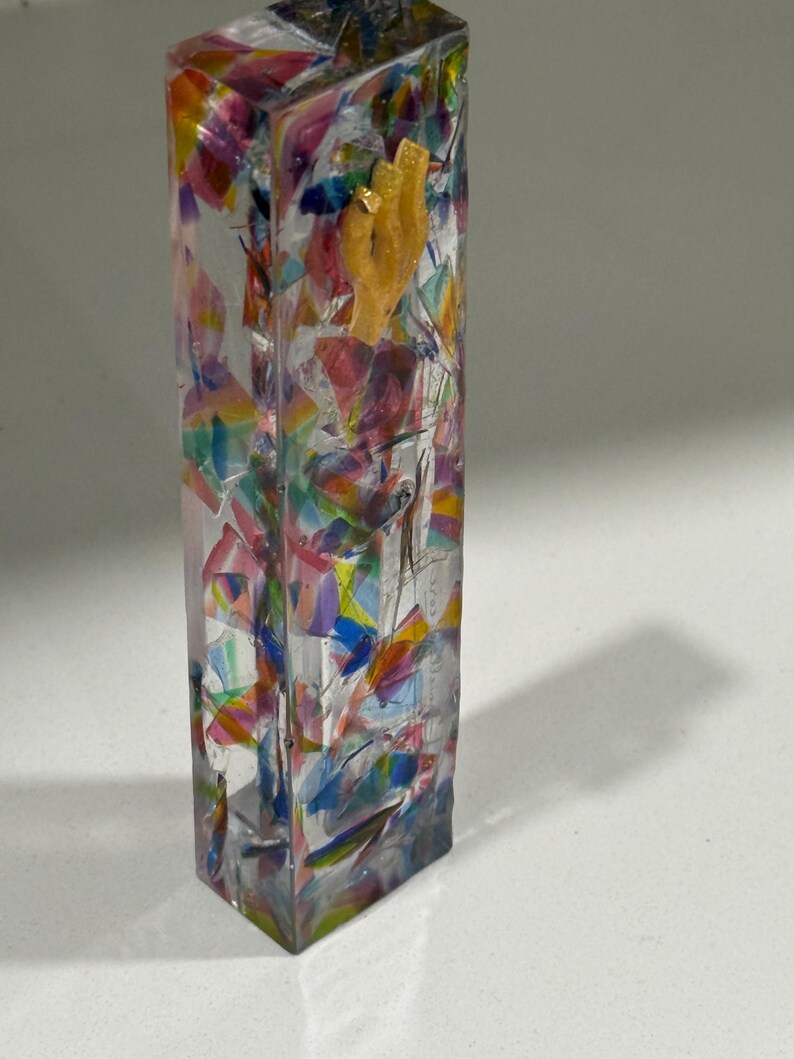 May include: A rectangular clear resin mezuzah case featuring an embedded mosaic of multicolored translucent shards in shades of pink, blue, yellow, and green, with a small, textured, light-colored Hebrew letter shin attached to the upper front surface.