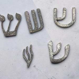 May include: Five distinct silver-colored Hebrew letter Shin shapes with a textured, glittery finish. The letters vary in size and calligraphic style, featuring curved and angular lines. These decorative pieces are arranged in a scattered layout.