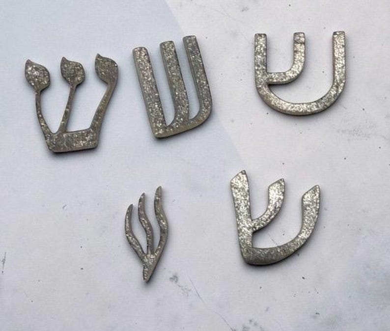 May include: Five distinct silver-colored Hebrew letter Shin shapes with a textured, glittery finish. The letters vary in size and calligraphic style, featuring curved and angular lines. These decorative pieces are arranged in a scattered layout.