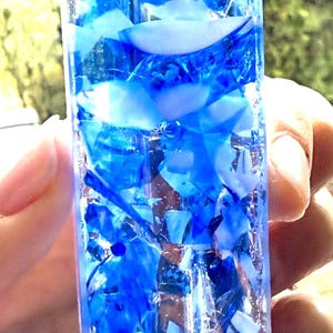 May include: A rectangular mezuzah case crafted from clear glass filled with a mosaic of blue and white glass shards. The top section features the black Hebrew letter Shin, which is clearly visible against the layered blue and white glass fragments.