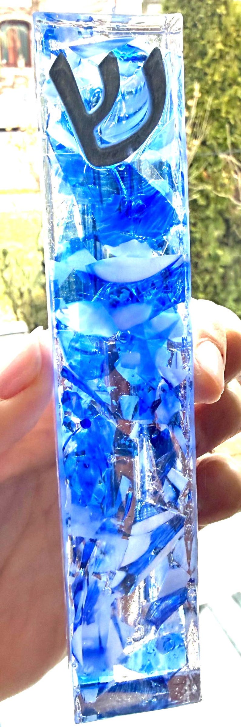 May include: A rectangular mezuzah case crafted from clear glass filled with a mosaic of blue and white glass shards. The top section features the black Hebrew letter Shin, which is clearly visible against the layered blue and white glass fragments.