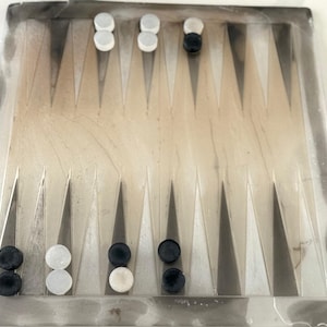 May include: A backgammon board with a light beige playing surface and dark gray border. The board features triangular points and is populated with white and black game pieces. The game pieces are arranged in a starting position, ready for play.