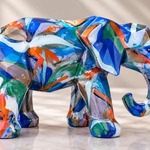 May include: A geometric elephant figurine featuring a multi-colored paint pour design with vibrant streaks of blue, orange, green, and white. The sculpture has a faceted, angular surface texture and stands in a side profile position.