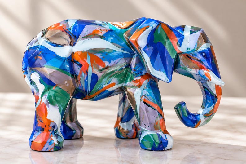 May include: A geometric elephant figurine featuring a multi-colored paint pour design with vibrant streaks of blue, orange, green, and white. The sculpture has a faceted, angular surface texture and stands in a side profile position.