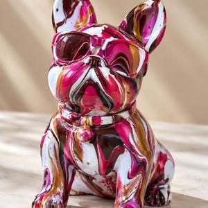 May include: A French bulldog figurine featuring a vibrant hydro-dipped paint pattern in shades of magenta, white, black, and gold. The dog is wearing sculpted sunglasses and a collar with a small round charm, all finished in a glossy sheen.