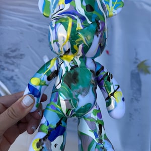 May include: A bear-shaped figurine featuring a glossy white base coat covered in a vibrant, multi-colored paint drip pattern. The surface displays swirling streaks of bright green, royal blue, yellow, and purple paint in a fluid, abstract design.