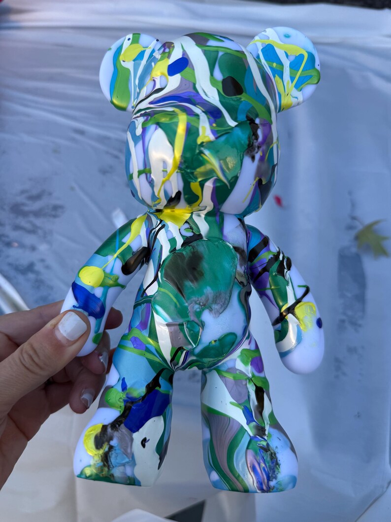 May include: A bear-shaped figurine featuring a glossy white base coat covered in a vibrant, multi-colored paint drip pattern. The surface displays swirling streaks of bright green, royal blue, yellow, and purple paint in a fluid, abstract design.