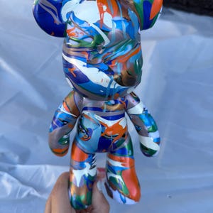 May include: A bear-shaped figurine featuring a glossy finish with a vibrant, multi-colored paint drip pattern in shades of blue, orange, white, green, and metallic bronze. The figure has rounded ears, a smooth surface, and a stylized humanoid form.