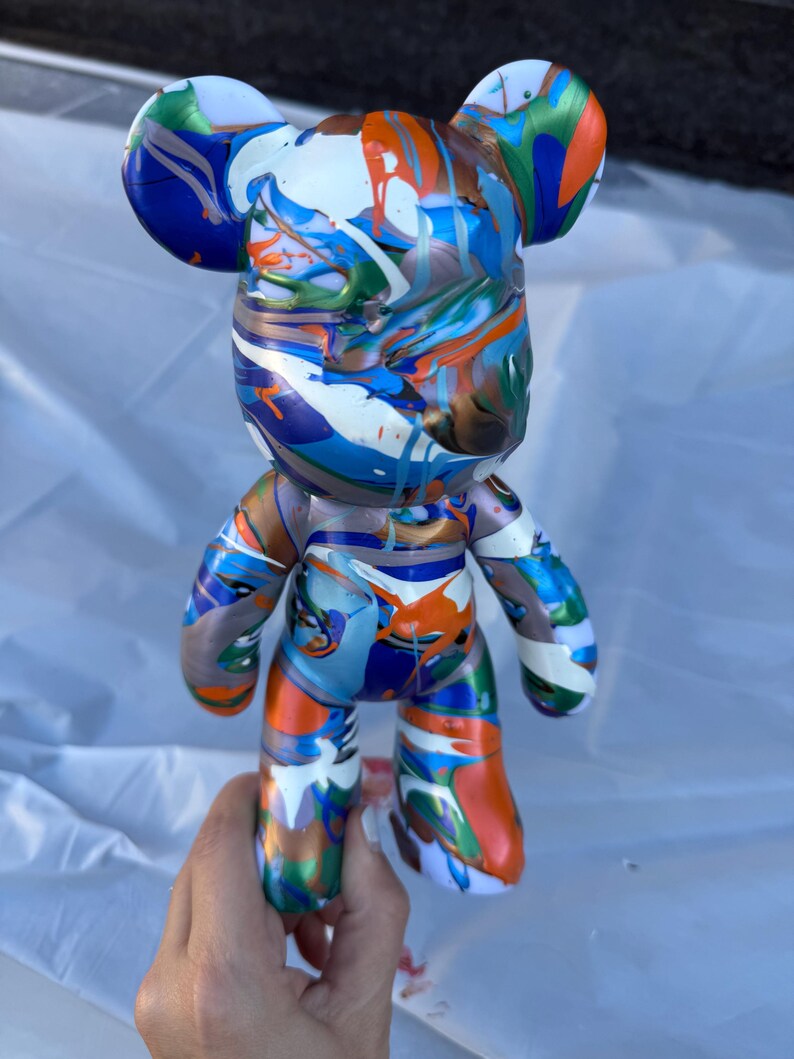May include: A bear-shaped figurine featuring a glossy finish with a vibrant, multi-colored paint drip pattern in shades of blue, orange, white, green, and metallic bronze. The figure has rounded ears, a smooth surface, and a stylized humanoid form.
