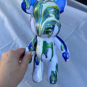 May include: A bear-shaped figurine with a white base color, featuring a fluid paint pour design in shades of blue, green, and yellow. The paint drips down from the head and body, creating a marbled, glossy effect across the surface.
